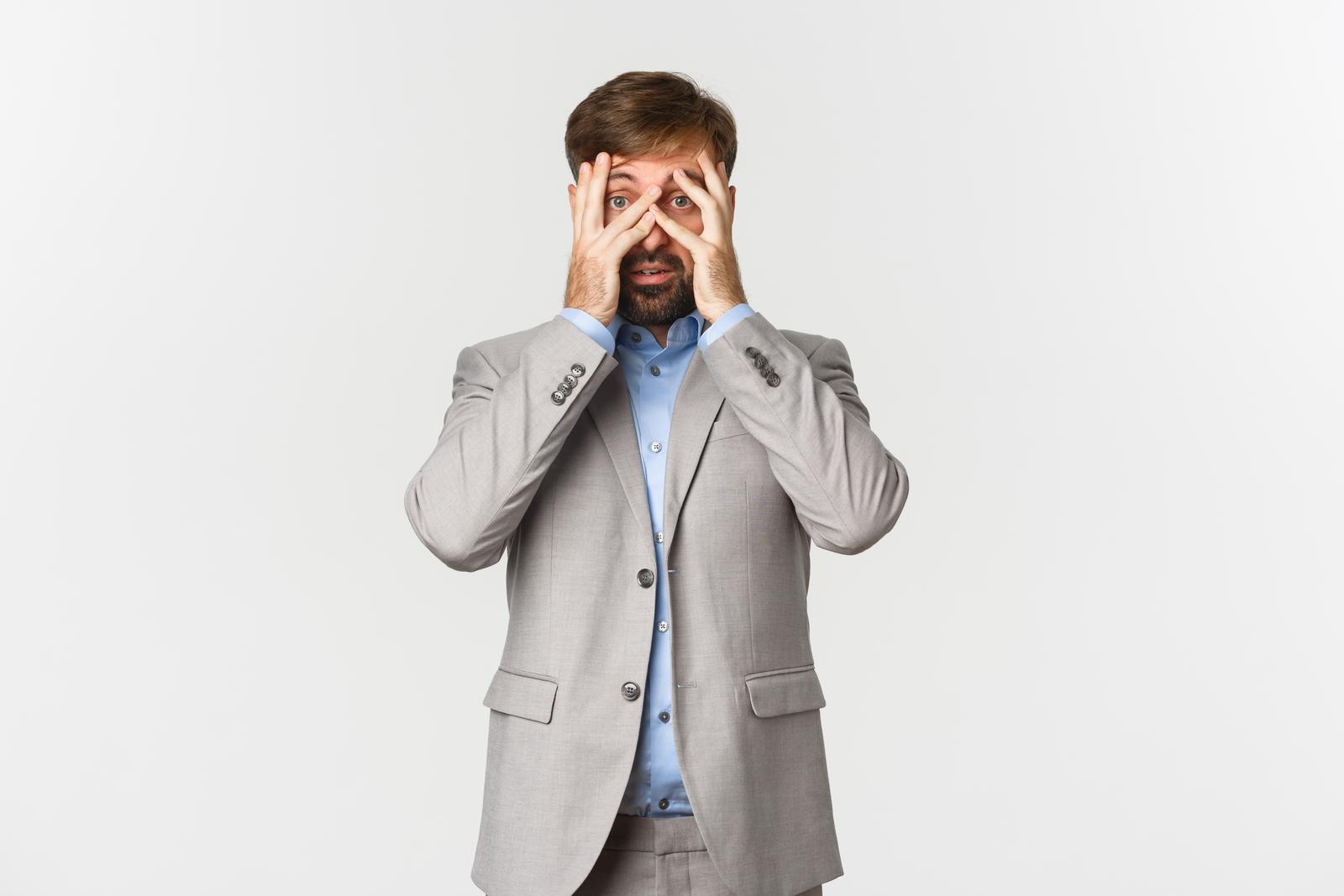 Portrait of businessman in grey suit, cover eyes and peeking through fingers with scared face.